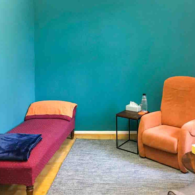 Der Behandlungsraum von Reiner Müller mit Coachingecke mit zwei Sesseln, einem runden Tisch und einer Hypnoseliege. Auf dem Tisch steht ein Blumenstrauß. Die Wand ist türkis gestrichen. Durch das Fenster scheint Licht herein.