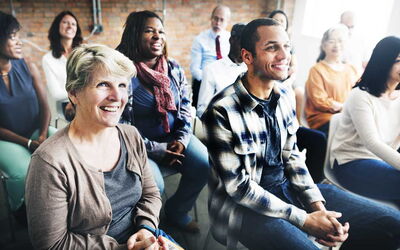 Ein Gruppe von Menschen sitzt als Publikum und schaut in eine Richtung zu einer Person, die etwas präsentiert und freut sich
