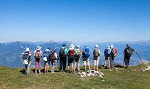 Eine Gruppe von Wanderern mit Rucksäcken steht im Gebirge an einem Hang und schaut ins Tal. Sie sehen entspannt aus.