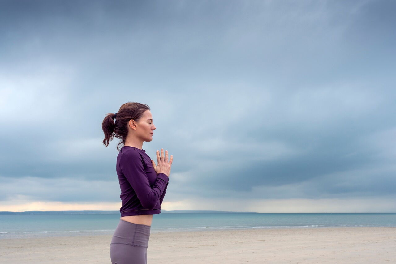 Eine junge Frau steht am Meer. Der Himmel ist wolkig und düster. Sie hat beide Hände vor der Brust verschränkt und die Augen geschlossen. Sie konzentriert sich auf ihren Atem, um zur Ruhe zu kommen.