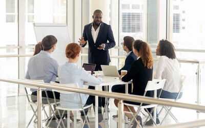 David steht im Anzug und hält eine Präsentation für eine kleine Gruppe von Menschen in einem modernen hellen Konferenzraum. Er steht vor einem Flipchart und spricht zu den Mitarbeiter*innen, die aufmerksam sind und Laptops oder Notizbücher vor sich haben.