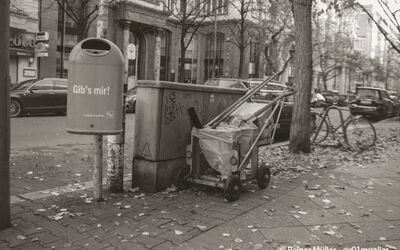 Ein Mülleimer auf der Straße. Daneben steht ein Wagen mit einem Besen.
