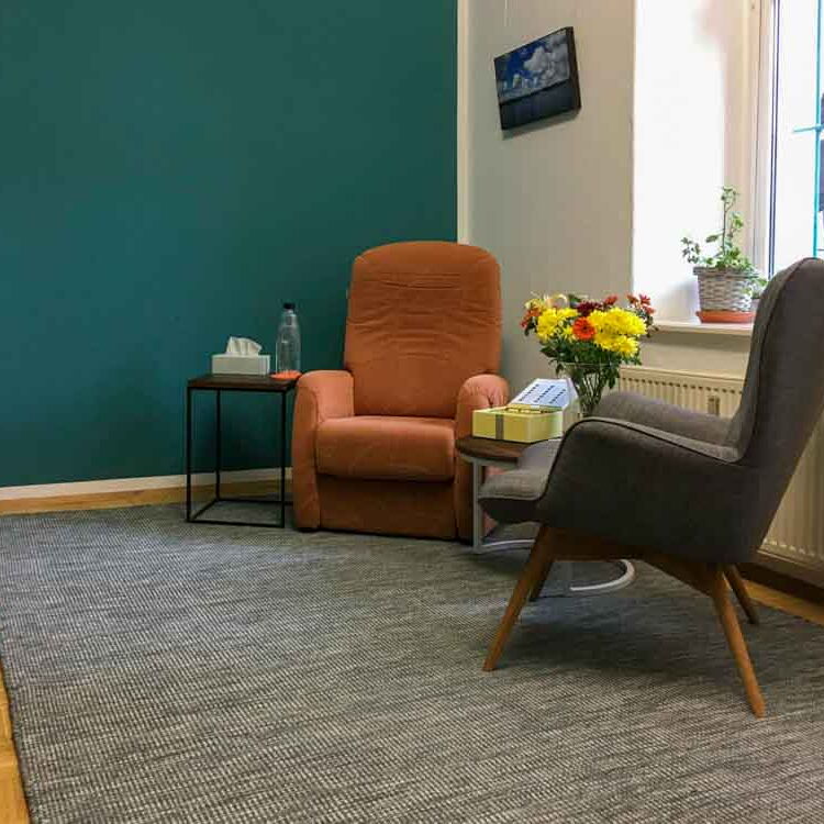 Der Behandlungsraum von Reiner Müller mit Coachingecke mit zwei Sesseln, einem runden Tisch und einer Hypnoseliege. Auf dem Tisch steht ein Blumenstrauß. Die Wand ist türkis gestrichen. Durch das Fenster scheint Licht herein.