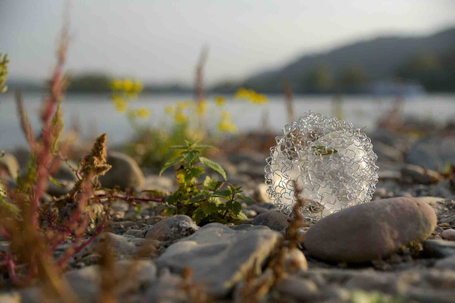 Eine zerbrochene Glaskugel, die von einem Drahtgeflecht umgeben ist, liegt am Ufer eines Flusses. Steine und einige Pflanzen sind in der Nähe der Kugel zu sehen.