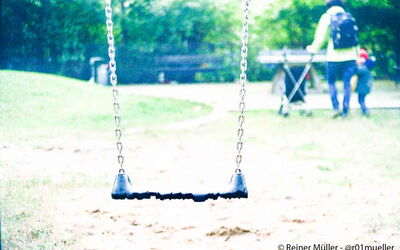 Eine leere Schaukel auf einem Spielplatz. Ein kleines Kind läuft an der Hand ihrer Mutter weg. Es schaut zurück zur Schaukel.
