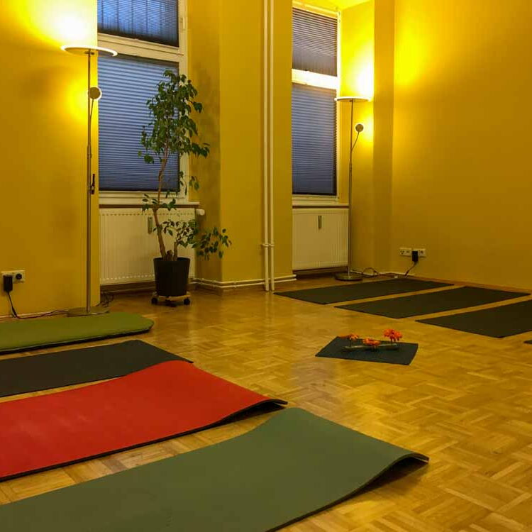 Der große Raum in der Coachingpraxis mit gelben Wänden, gedämpften Licht und zugezogenen Rollos. Auf dem Boden liegen Yoga-Matten.