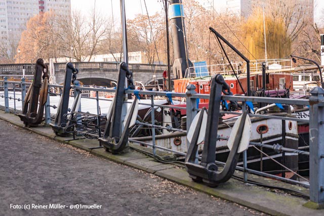 An einer Hafenmole liegen alte Dampfer. Am Ufer stehen vier alte Anker.