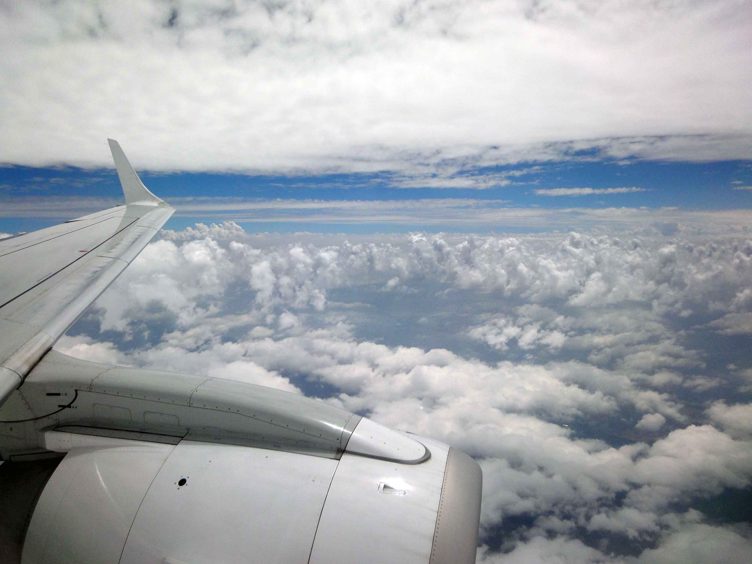 Der Blick aus dem Fenster eines Flugzeuges auf die Tragflächen und einen wolkenreichen und sonnigen Himmel. Unbeschwert ohne Flugangst in den Urlaub fliegen, wir unterstützen Sie mit Hypnose dabei.
