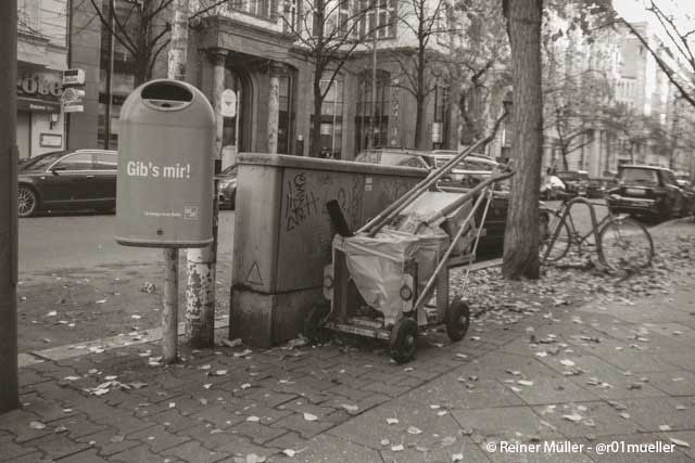 Ein Mülleimer auf der Straße. Daneben steht ein Wagen mit einem Besen.